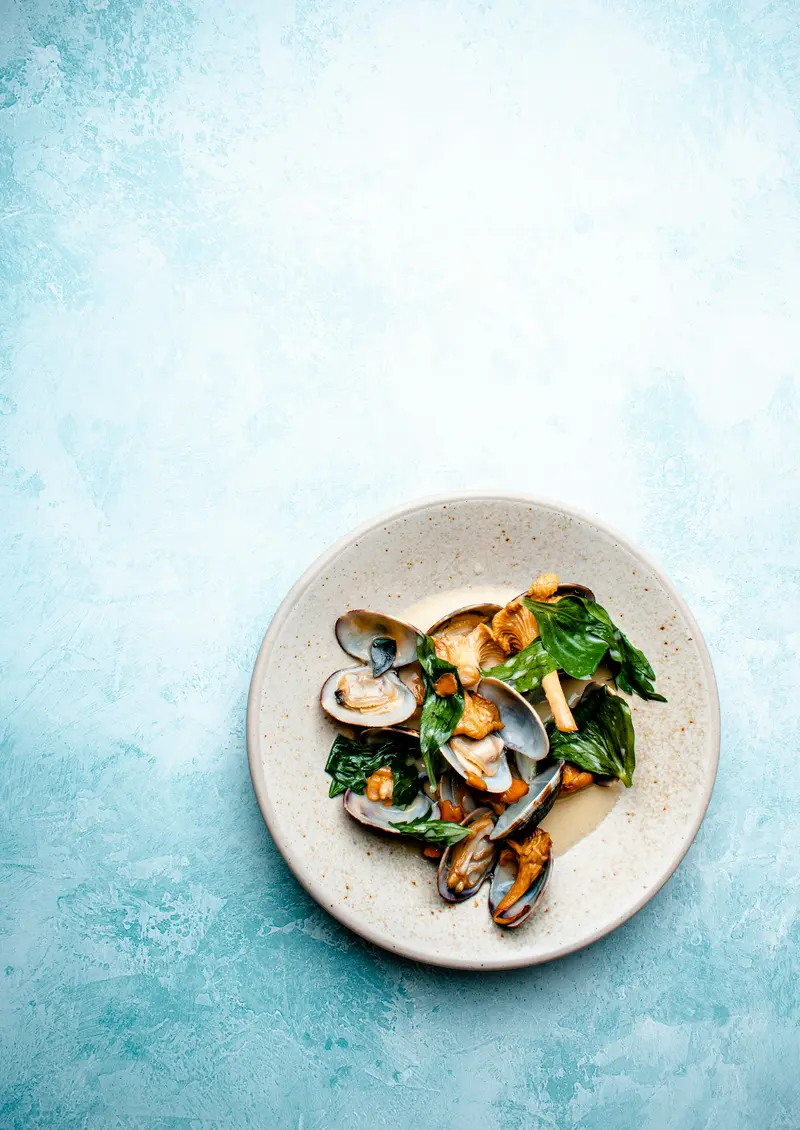 A bowl of clams with greens on a pale blue table